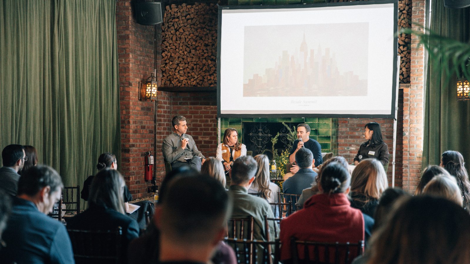 Four panelists speaking on stage in front of a brick fireplace and a projection screen during the Beyond Year One panel, with the audience seated in the foreground