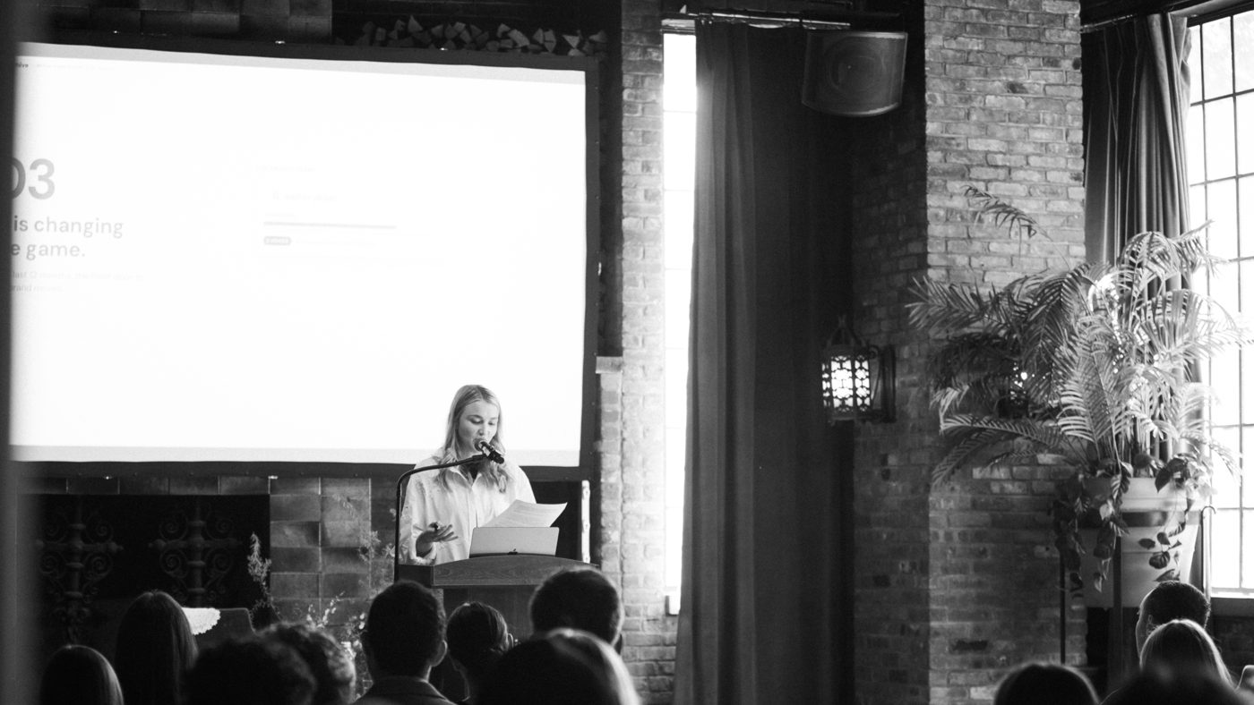 Black-and-white photo of Emily Gittins speaking at a podium in front of a projected slide, with the audience seated in the foreground