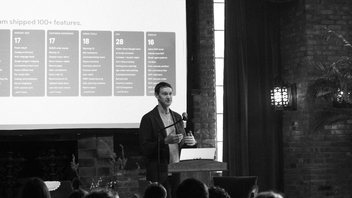 Black-and-white photo of Ryan Rowe speaking at a podium in front of a slide reading "shipped 100+ features," with the audience seated in the foreground